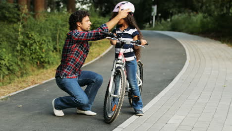 Closeup.-Portrait-of-a-pretty-girl-and-her-father-near-the-bike.-Dad-wears-a-helmet-on-the-girl's-head.-Smiling.-Blurred-background